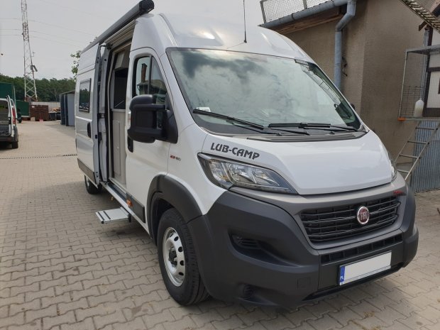 Biały kamper Fiat z otwartymi drzwiami bocznymi i napisem LUB-CAMP, zaparkowany na placu.