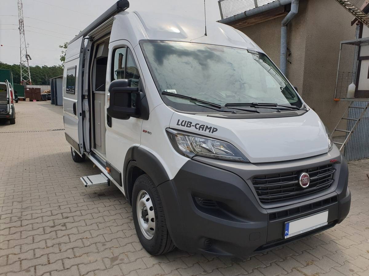 Biały kamper Fiat z otwartymi drzwiami bocznymi i napisem LUB-CAMP, zaparkowany na placu.