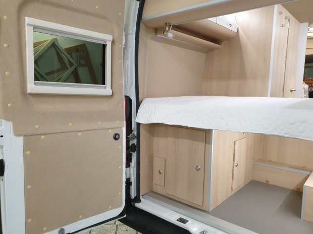 Wnętrze kampera: łóżko nad szafkami z jasnego drewna i otwarte tylne drzwi