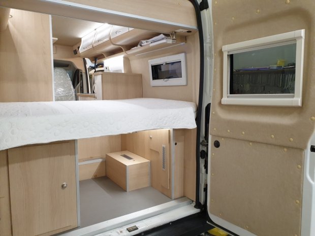 Wnętrze kampera z łóżkiem, jasnymi drewnianymi szafkami i małym okienkiem.