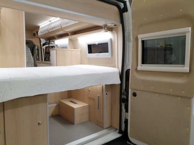Wnętrze kampera: łóżko, jasne szafki i okno w drzwiach
