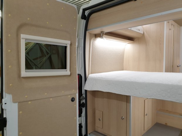 Wnętrze kampera: łóżko z białym materacem, drewniane szafki i otwarte tylne drzwi z oknem