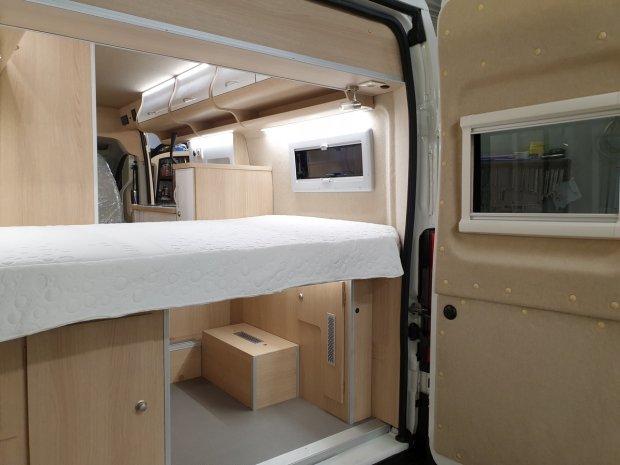 Wnętrze kampera z łóżkiem, drewnianymi szafkami i okienkiem w bocznych drzwiach