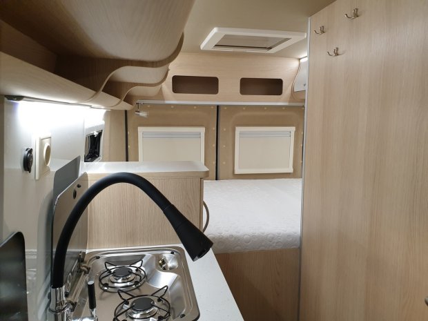 Wnętrze kampera: mała kuchnia z dwupalnikowym palnikiem i łóżkiem w jasnym drewnie