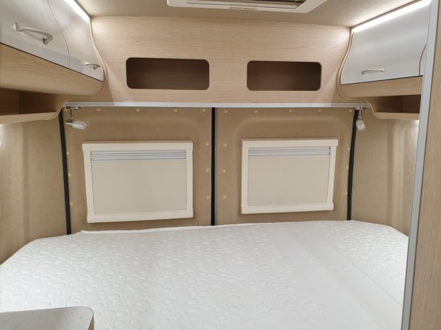 Wnętrze kampera: łóżko z białym materacem, zabudowa drewniana i rolety przy oknach