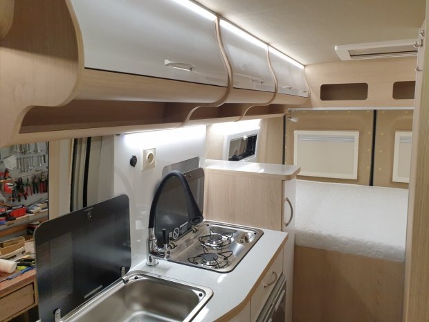 Wnętrze kampera z kuchnią: zlew, bateria, płyta gazowa i łóżko w tle.