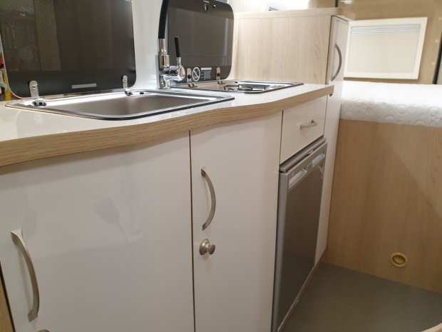 Kompaktowa kuchnia w kamperze: zlew, płyta gazowa, białe szafki i mała lodówka