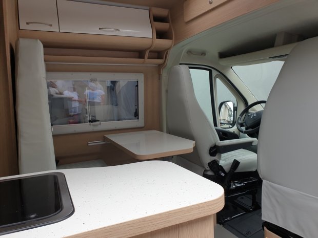 Wnętrze kampera z białym blatem, stolikiem i obróconymi przednimi fotelami.