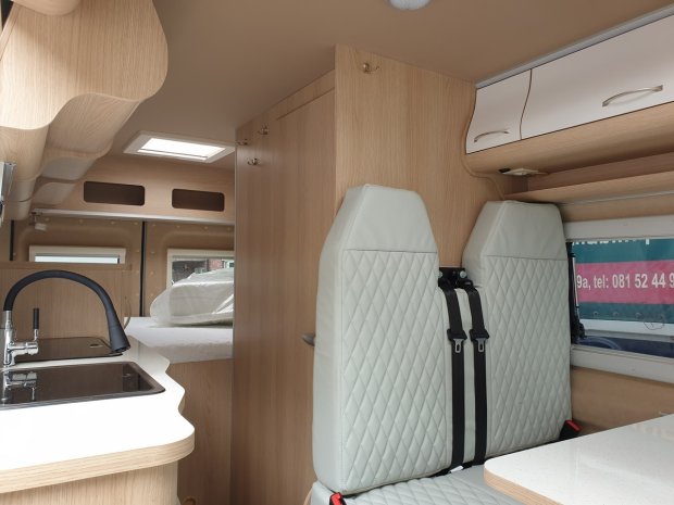 Wnętrze kampera z pikowanymi jasnymi fotelami, drewnianymi szafkami, zlewem.