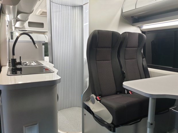 Wnętrze kampera z kuchenką, zlewem i dwoma ciemnymi fotelami przy stoliku