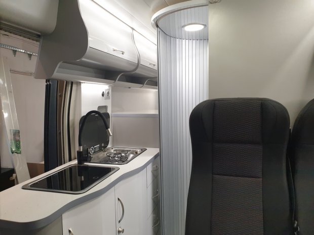 Wnętrze kampera: aneks kuchenny z umywalką, kuchenką, szafkami i fotelem.