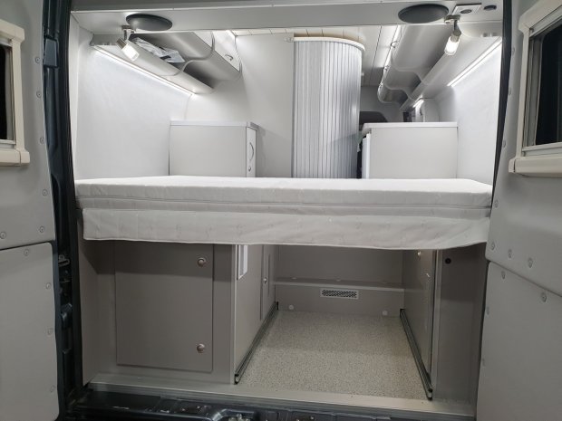 Wnętrze kampera z łóżkiem na podwyższeniu i szafkami, jasnoszare.