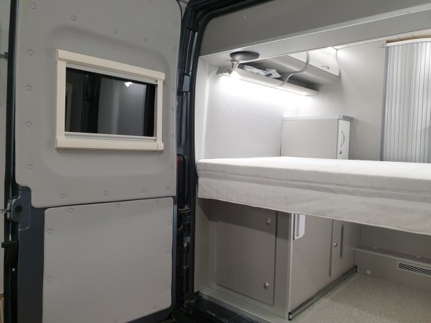 Wnętrze kampera: rozkładane łóżko, półki nad nim i małe okno w drzwiach.