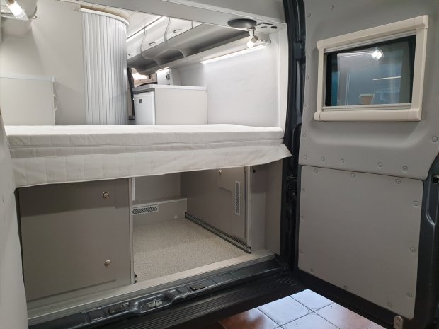 Wnętrze kampera: łóżko nad schowkami, mini lodówka i małe okno w drzwiach.