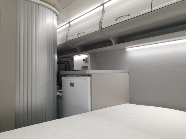 Wnętrze kampera: łóżko, lodówka, szafki nad głową i przesuwna żaluzja.