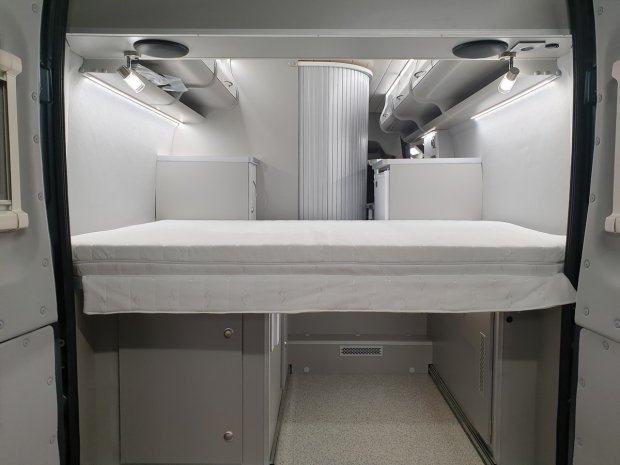 Wnętrze kampera z łóżkiem, białymi szafkami i oświetleniem LED