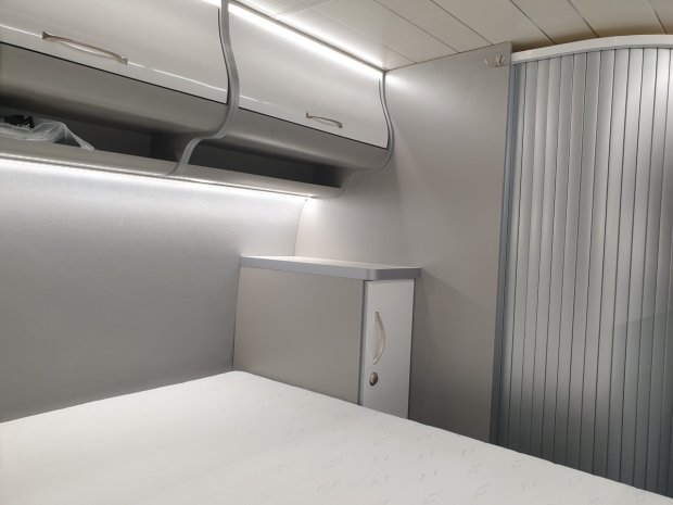 Wnętrze przytulnej kamperowej sypialni z łóżkiem, szafką i podświetlanymi półkami.