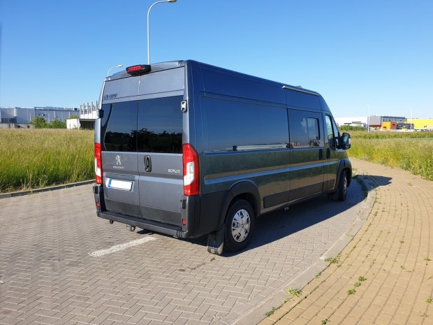 Ciemnoszary Peugeot Boxer zaparkowany bokiem i tyłem na brukowanym parkingu przy polu.