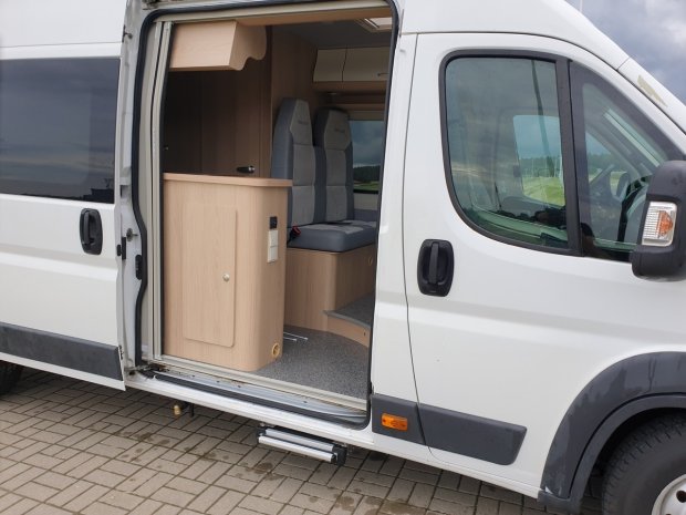 Biały kamper z otwartymi drzwiami, widoczne drewniane meble i siedzenia.