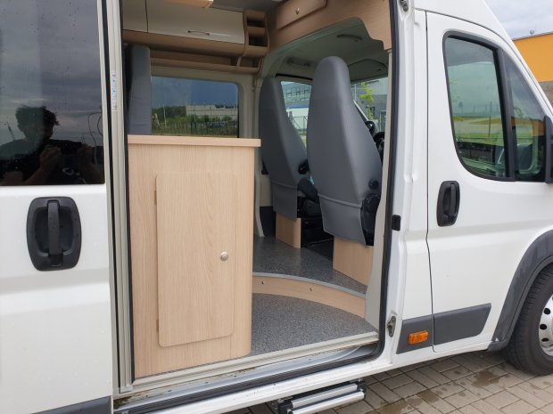 Otwarty biały kamper, drewniana szafka przy wejściu i dwa szare fotele.