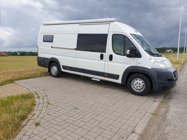 Biały kamper dostawczy zaparkowany na chodniku obok pola pod pochmurnym niebem
