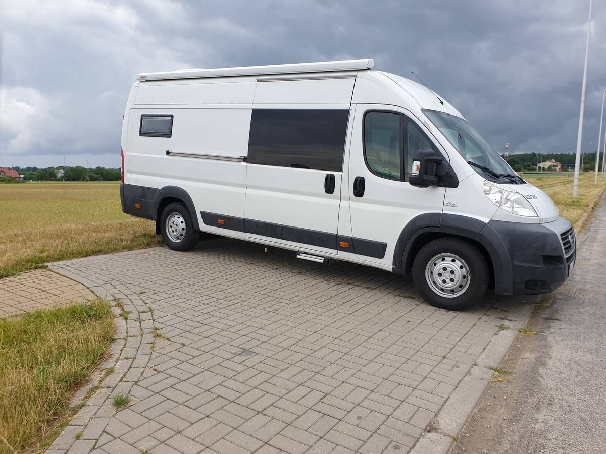 Biały kamper dostawczy zaparkowany na chodniku obok pola pod pochmurnym niebem