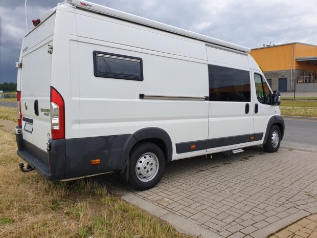 Biały bus-kamper zaparkowany przy chodniku przed pomarańczową halą.