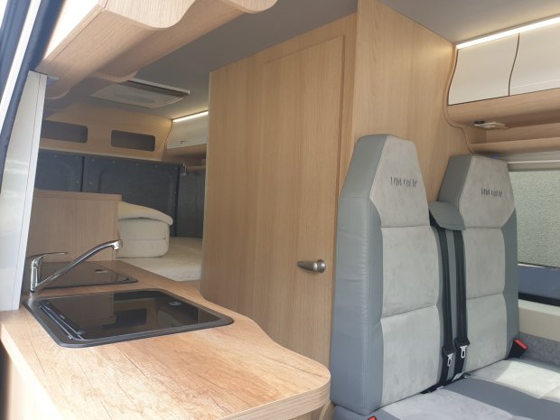 Wnętrze kampera: drewniana kuchnia ze zlewem, szafkami, dwoma szarymi fotelami i łóżkiem.