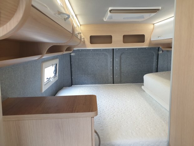 Wnętrze kampera: łóżko z białym materacem, drewniane szafki nad głową i małe okienko