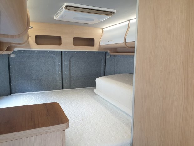 Aranżacja sypialni w kamperze: łóżko, jasne drewniane szafki i otwór wentylacyjny.