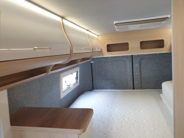 Wnętrze kampera: łóżko, szafki górne z oświetleniem i małe okno.