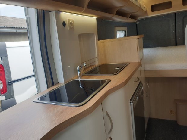 Wnętrze kampera z drewnianym blatem, zlewem i płytą kuchenną oraz łóżkiem.