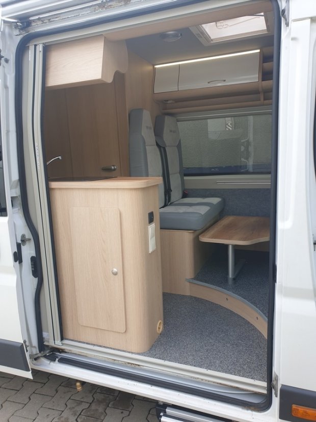Wnętrze kampera: jasne drewniane szafki, kanapa z pasami i składany stolik.