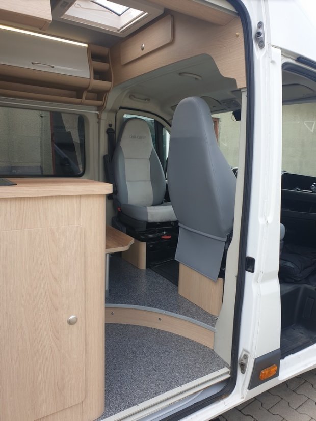 Wnętrze kampera: szare fotele kierowcy, jasne drewniane meble i podłoga.