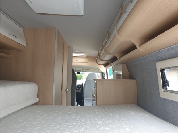 Wnętrze kampera: łóżko z materacem, szafki z jasnego drewna i przednie siedzenia.