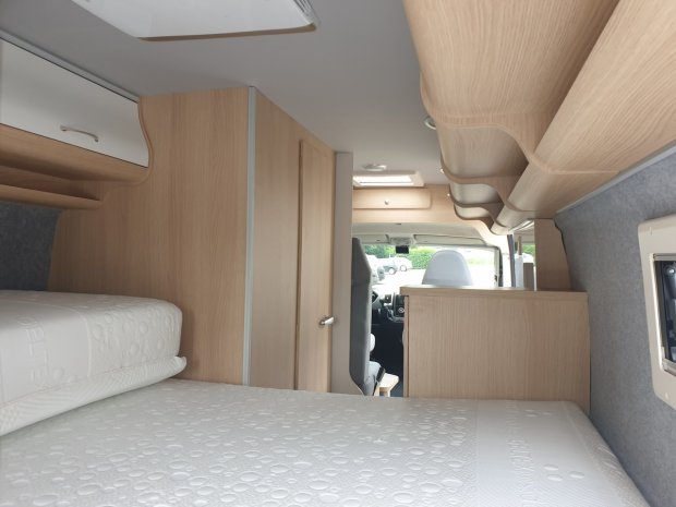 Wnętrze kampera: łóżko, jasne drewniane szafki i widok na kabinę kierowcy