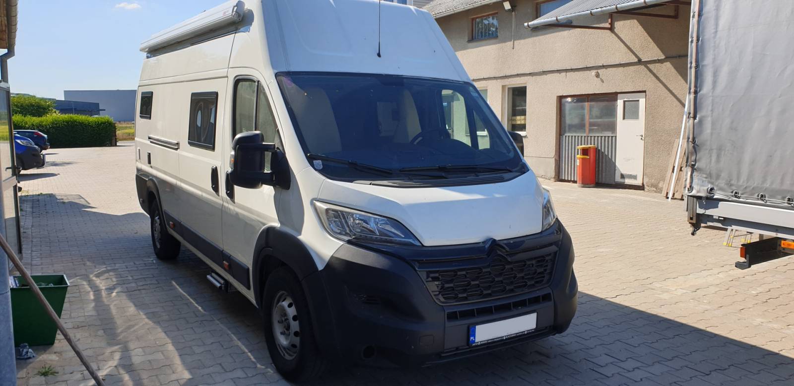 Biały van kempingowy zaparkowany na podwórzu obok budynku