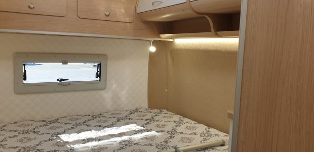 Łóżko w kamperze z wzorzystym materacem, małym oknem, drewnianymi szafkami i lampką.