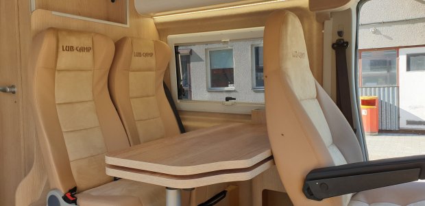 Wnętrze kampera: beżowe fotele z napisem LUB-CAMP i drewniany stolik między nimi