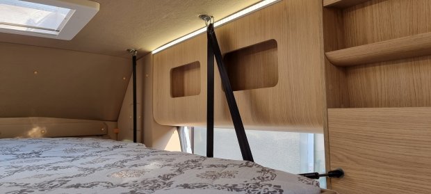 Przestrzeń sypialna kampera: łóżko z wzorzystą pościelą, drewniane półki i pasy zabezpieczające