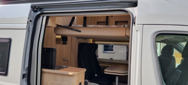 Wnętrze kampera przez drzwi: drewniane meble, opuszczane łóżko, fotele i stół.