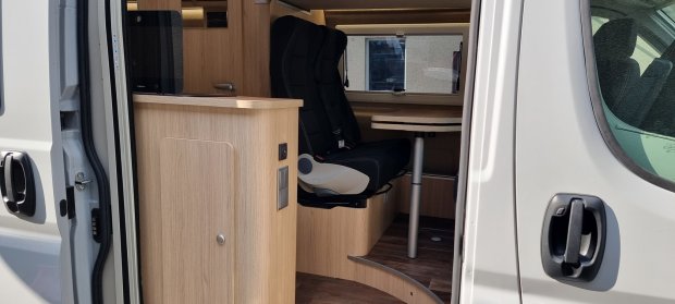 Wnętrze kampera przez otwarte drzwi: drewniane meble, obracane czarne siedzisko i stolik