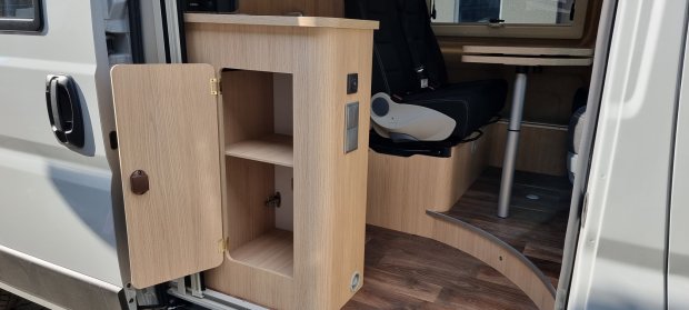 Wnętrze kampera z otwartą drewnianą szafką i obrotowym fotelem przy stoliku