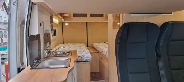 Wnętrze kampera: kuchnia z zlewem i płytą, dwa łóżka i przednie fotele.