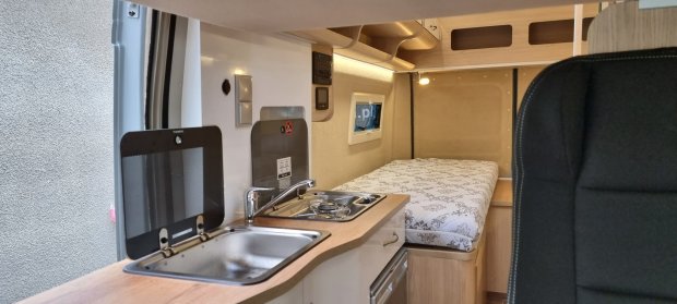 Wnętrze kampera: aneks kuchenny ze zlewem i kuchenką oraz łóżko z wzorzystym materacem.