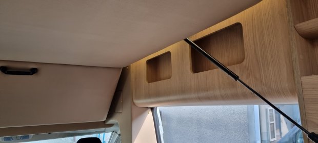 Wnętrze kampera: drewniana półka nad kabiną z otwartymi schowkami i amortyzatorem.