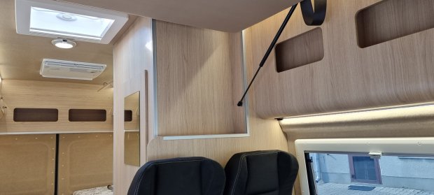 Wnętrze kampera z jasnym drewnem, oknem, świetlikiem i dwoma czarnymi fotelami