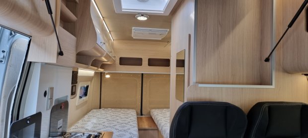 Wnętrze kampera z dwiema łóżkami, jasnymi drewnianymi szafkami i małą kuchnią.