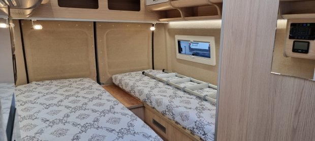 Wnętrze kampera z dwiema dostawnymi łóżkami, wzorzystymi materacami i składalną drabinką.