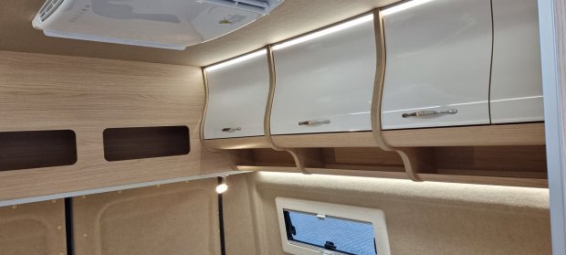 Wnętrze kampera z drewnianymi półkami, białymi szafkami i podświetleniem LED